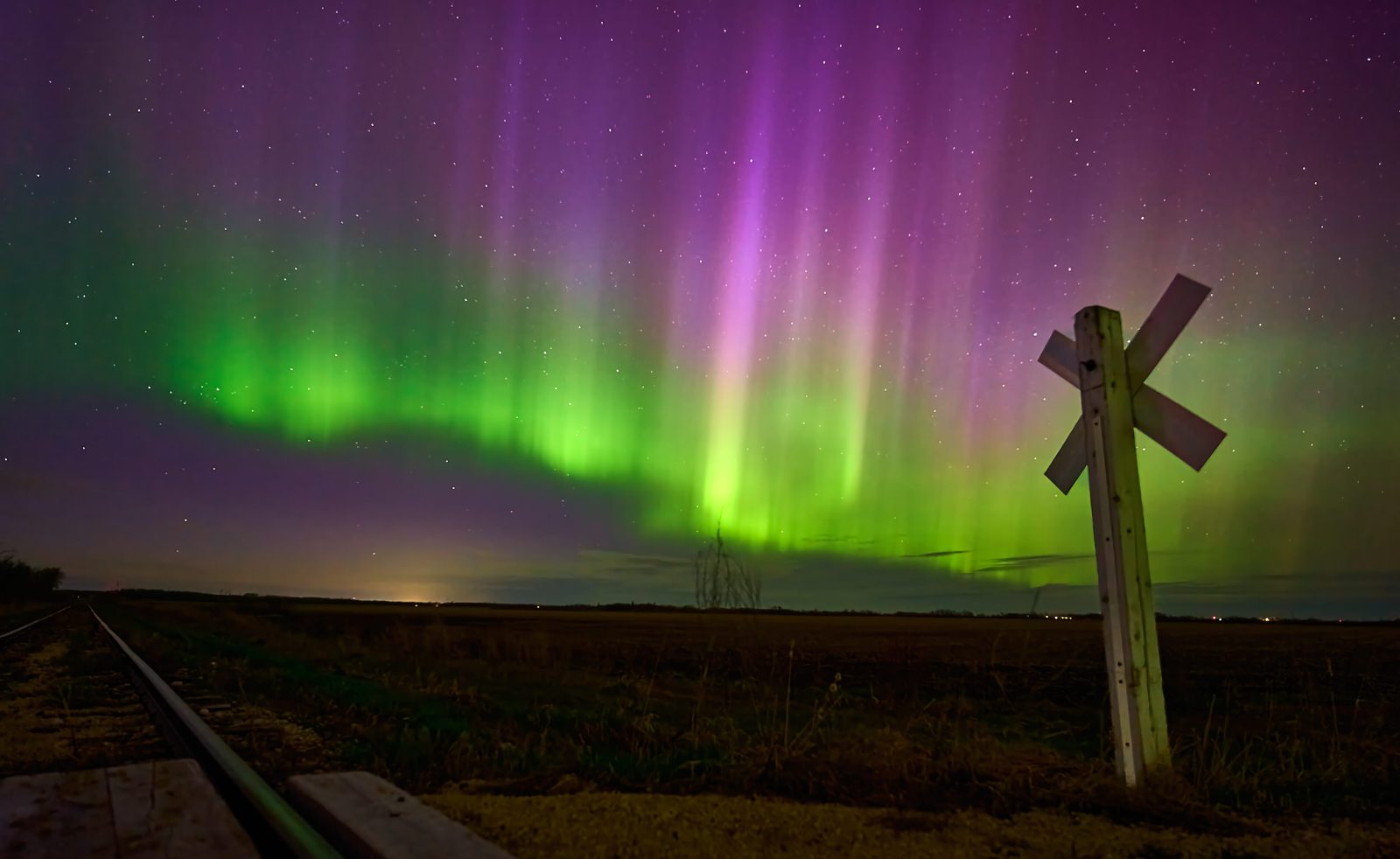 Purple pillars and a green arc of aurora over a prairie field with a railway crossing sign