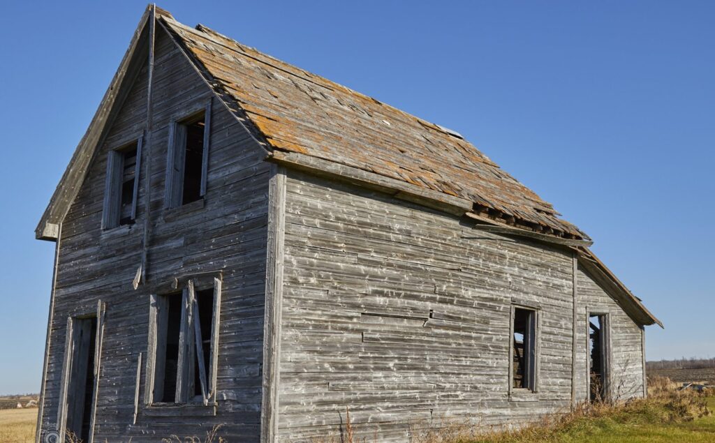 Weathered grey farmhouse with empty windows under a blue prairie sky