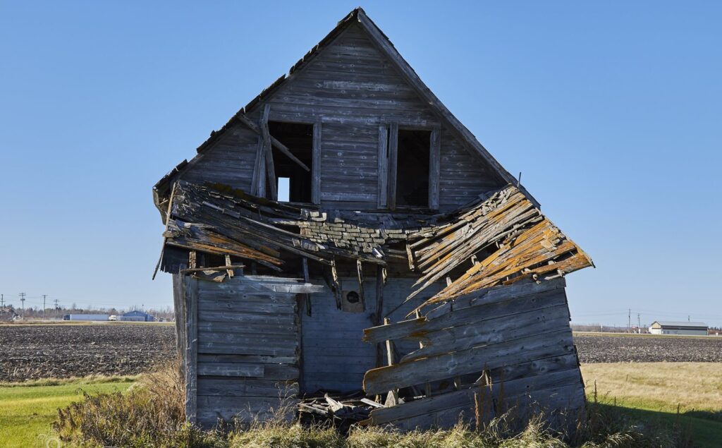 Leaning abandoned farmhouse with a collapsed roof beside a ploughed field