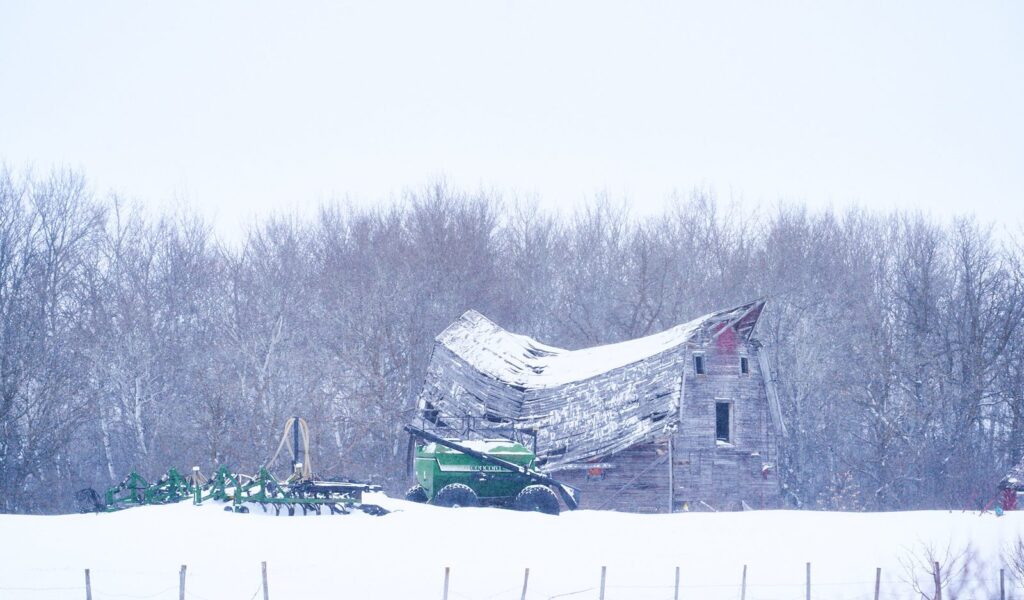 Collapsing barn in snow beside farm machinery