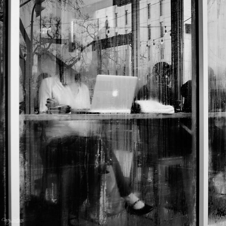 Two people working at a laptop seen through a rain-streaked café window