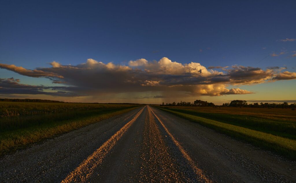 Gravel road with a yellow centre line stretching to the horizon under a cloud lit by evening sun