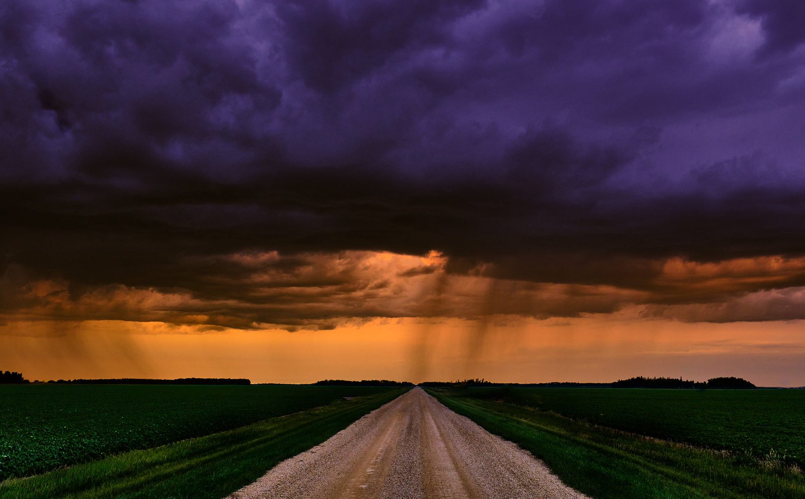 Gravel road leading toward a purple storm cloud with rain shafts and an orange horizon