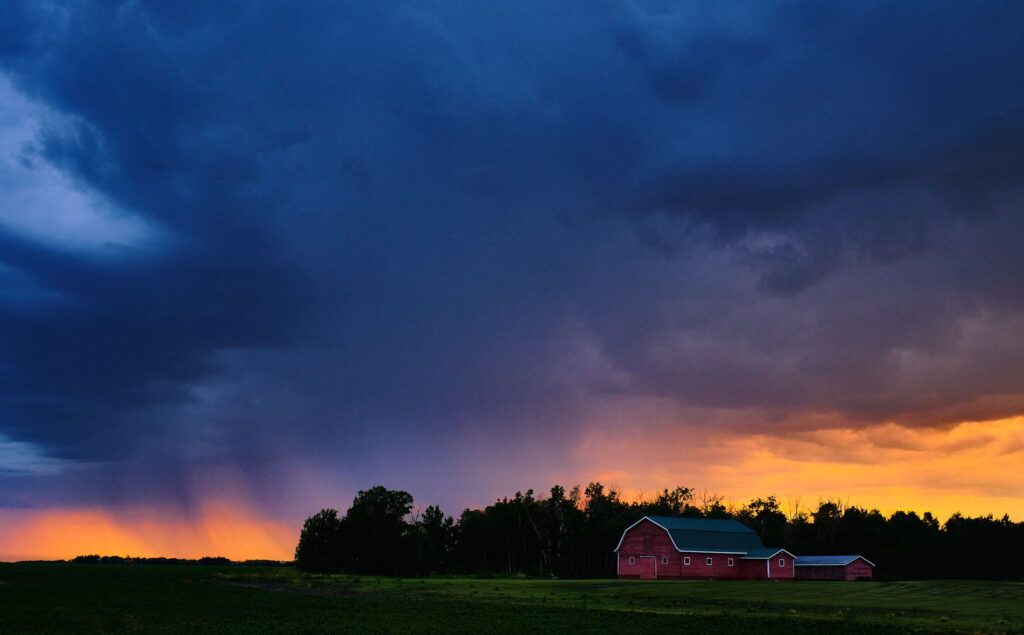 Red barn on the horizon beneath a blue storm cloud with rain and an orange sunset