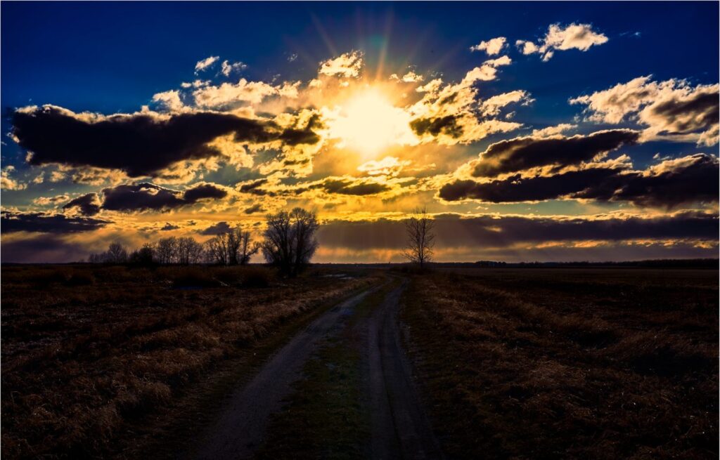 Sun starburst through broken cloud above a dirt track across a field