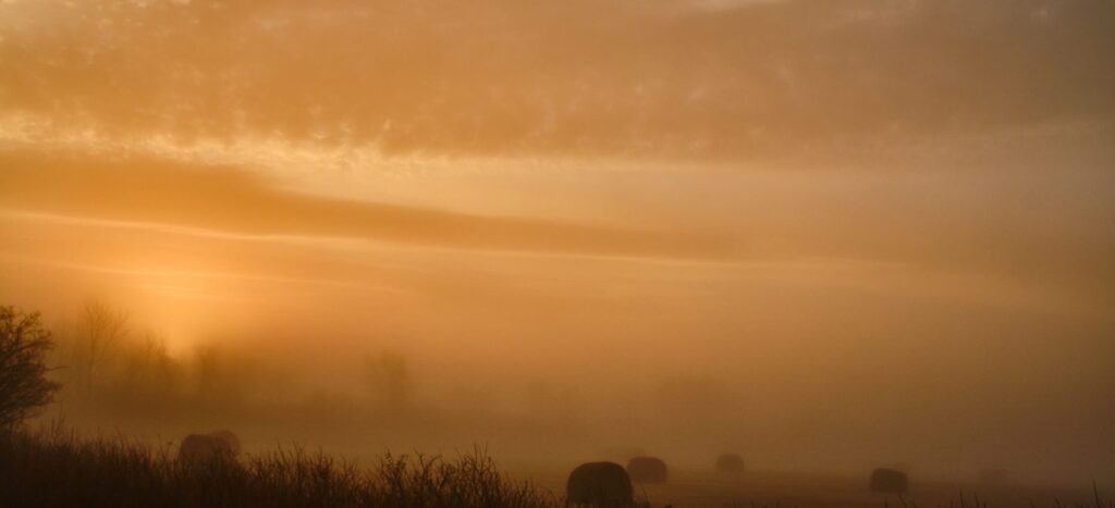 Orange sunrise glowing through fog over a prairie field with hay bales