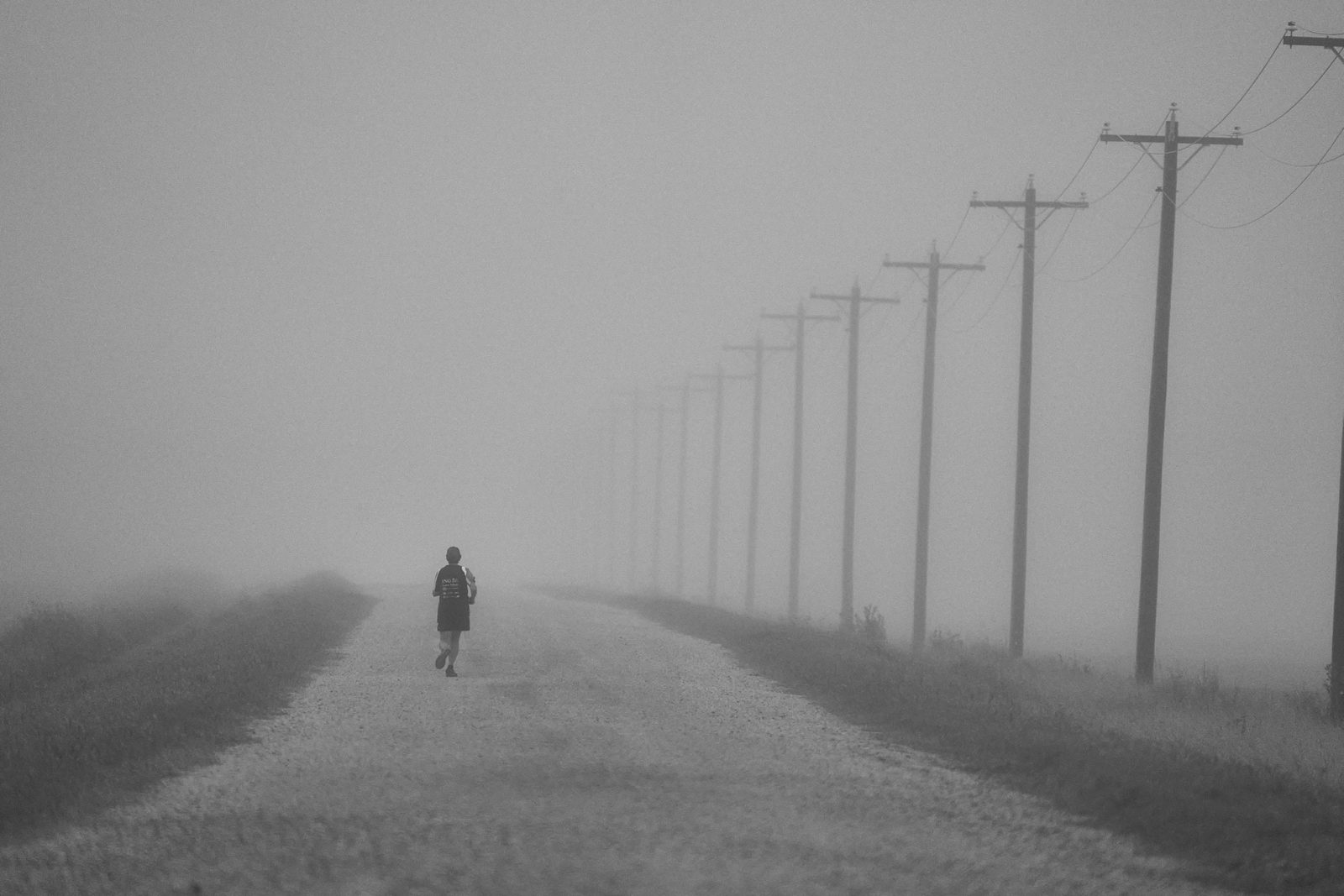 Person walking down a gravel road beside a line of power poles fading into fog
