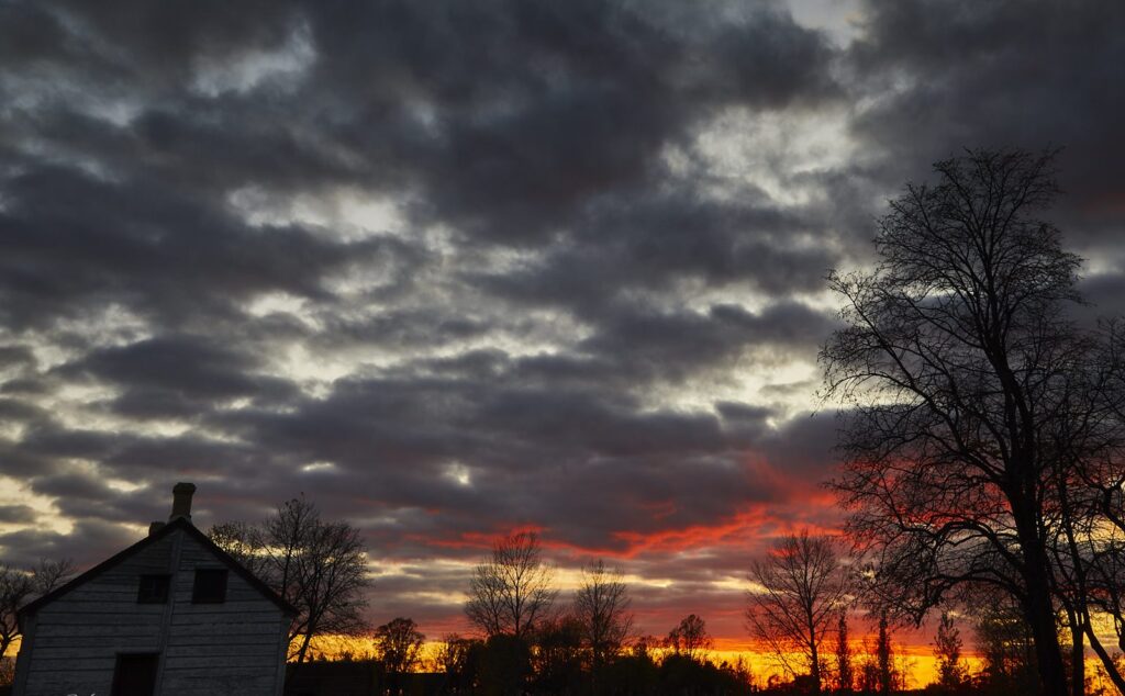 Dark clouds and red sunset behind a farmhouse and a bare tree