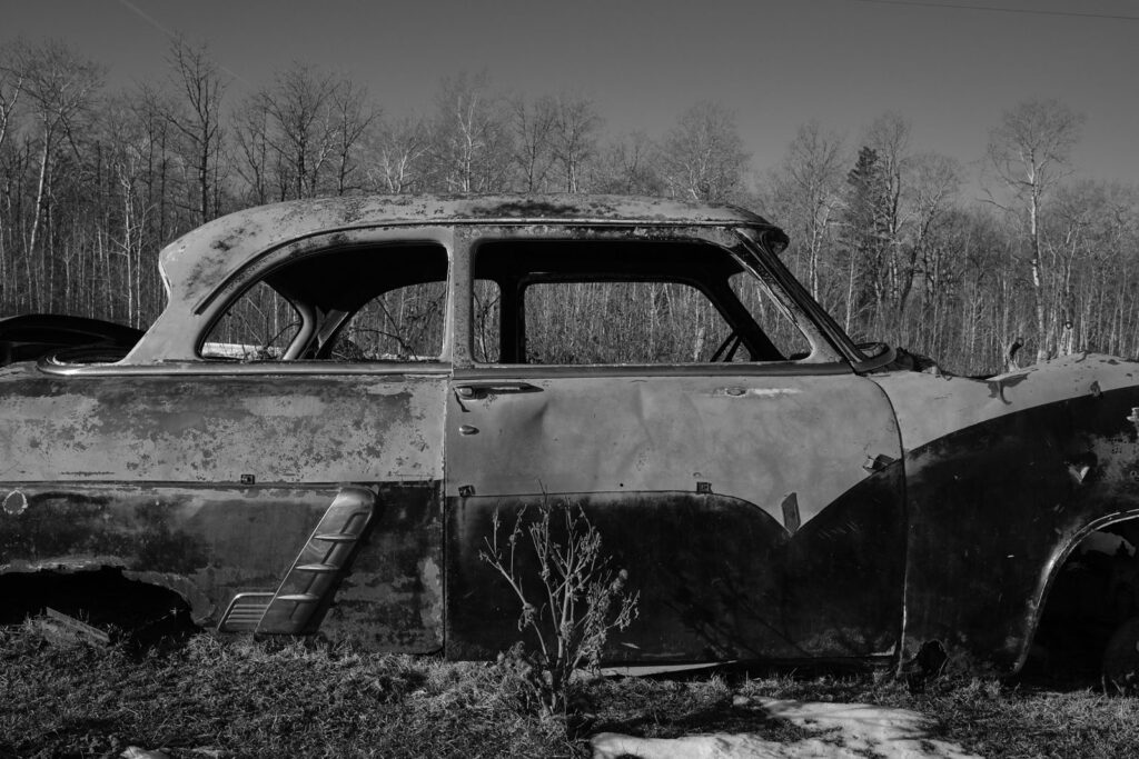 Burned-out vintage car body in a clearing with bare trees behind