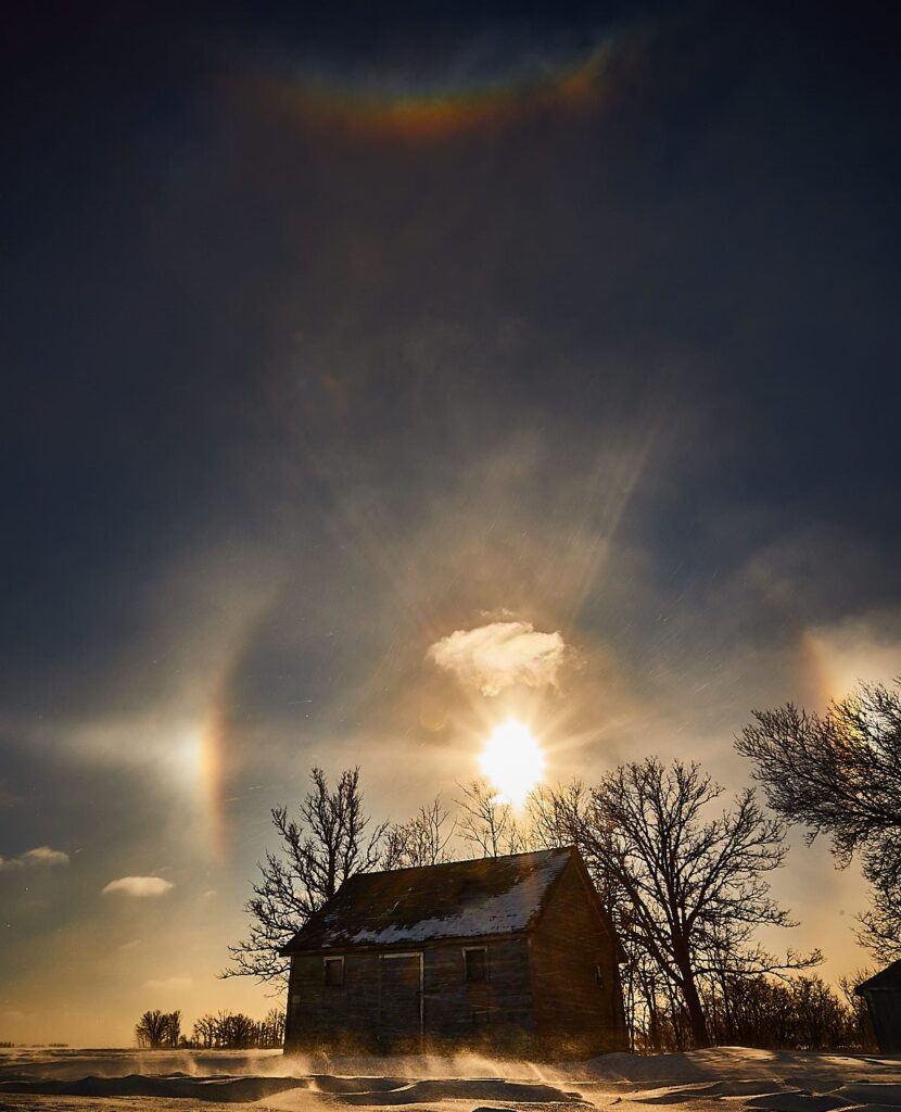 Sun dogs and halo glowing around the sun behind an old farmhouse in blowing snow